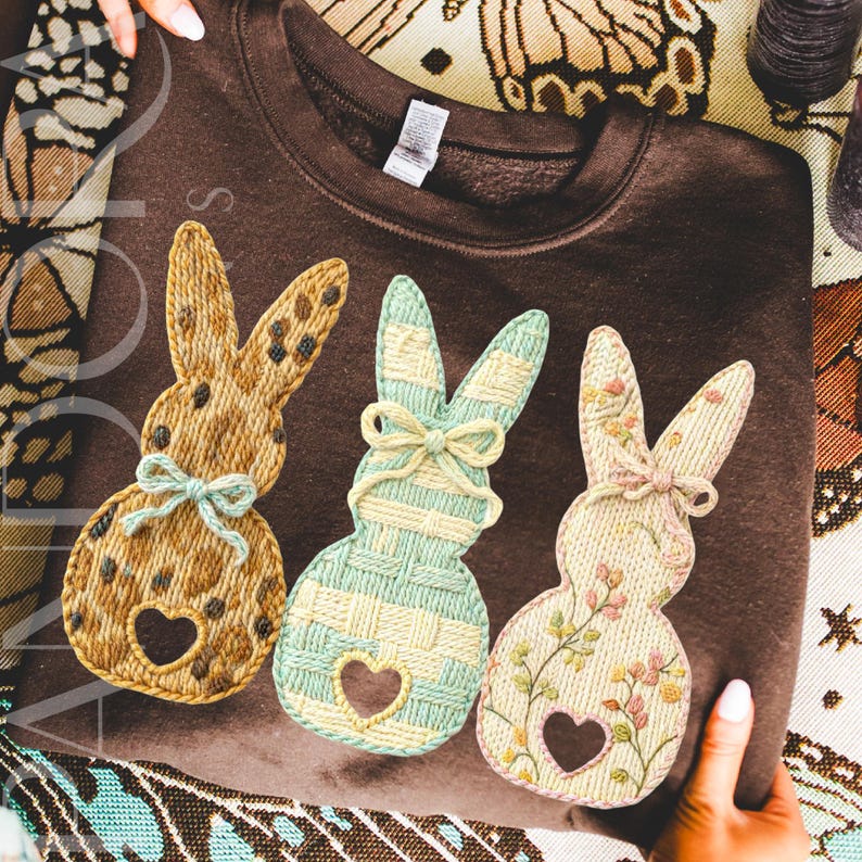 May include: A brown sweatshirt featuring three embroidered bunny designs. Each bunny has a heart-shaped cutout and a bow. The bunnies are in shades of tan, mint green, and pink, with floral and patterned details. The sweatshirt is a cozy, casual garment.