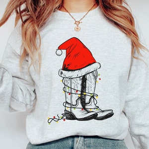 Pu&ograve; includere: Una felpa grigia con un disegno bianco di stivali da cowboy che indossano un cappello di Babbo Natale e luci di Natale.