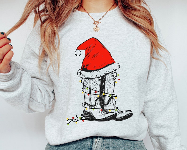 Pu&ograve; includere: Una felpa grigia con un disegno bianco di stivali da cowboy che indossano un cappello di Babbo Natale e luci di Natale.
