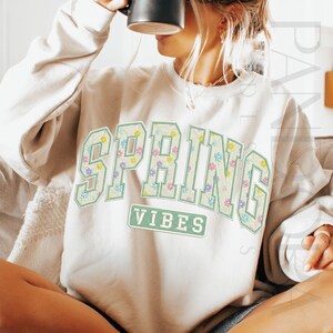 May include: Cream-colored sweatshirt with the words "SPRING VIBES" in a floral design. The letters are outlined in light green, with a floral pattern inside. The word "VIBES" is in a rectangular box below the word "SPRING".