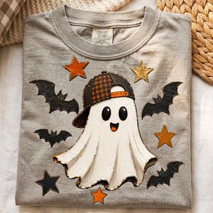 May include: A grey t-shirt featuring a central graphic of a white ghost wearing a black and orange plaid baseball cap. The design includes black bats and orange and gold stars surrounding the ghost. The shirt has a Comfort Colors brand label.