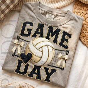 May include: A light gray t-shirt featuring the text GAME DAY in black block letters with gold outlines. A white volleyball graphic is centered between the words, accompanied by a black heart and two decorative gold bows over a net pattern.