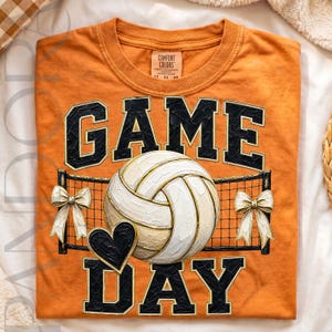 May include: An orange t-shirt featuring the text GAME DAY in black block letters with gold outlines. A white volleyball is centered in front of a black net graphic, flanked by two white bows and a small black heart icon below the ball.