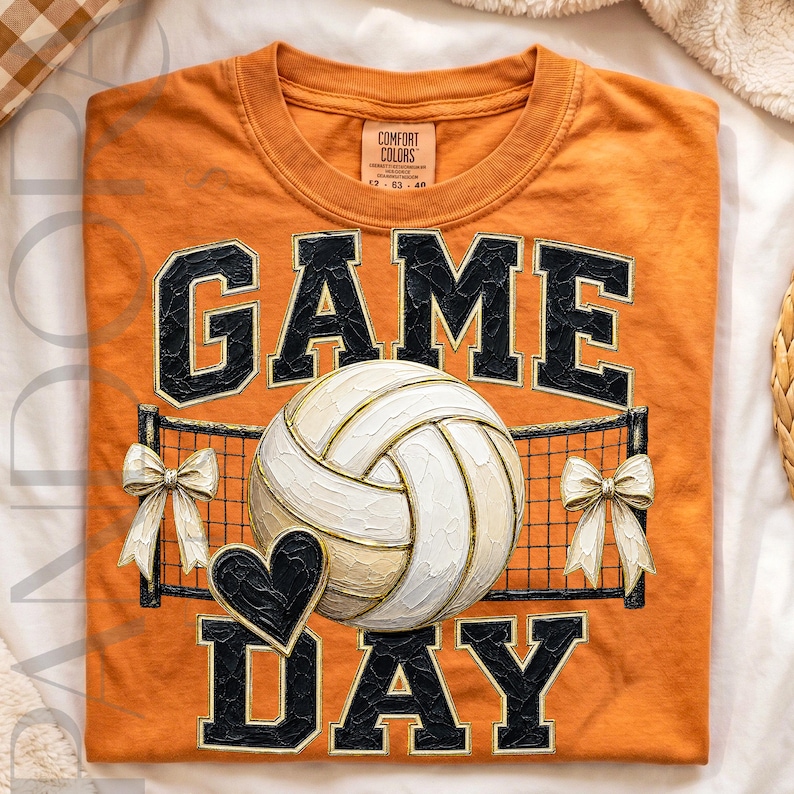 May include: An orange t-shirt featuring the text GAME DAY in black block letters with gold outlines. A white volleyball is centered in front of a black net graphic, flanked by two white bows and a small black heart icon below the ball.