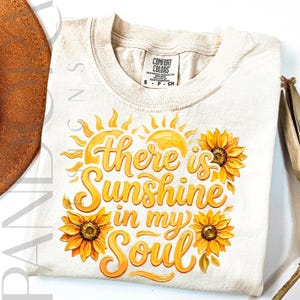 May include: Cream-colored t-shirt with the phrase "there is sunshine in my soul" in yellow script, accented by yellow sunflowers and sun graphics. The shirt is a Comfort Colors brand, size small.