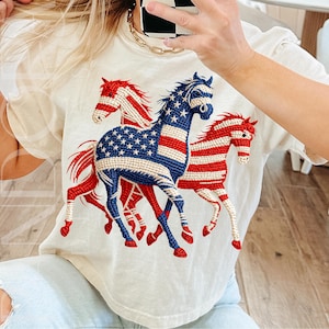 May include: Cream-colored t-shirt featuring a graphic of three running horses. The horses are decorated with red and white stripes and blue with white stars, resembling the American flag. The design is centered on the shirt.