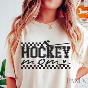 May include: A white t-shirt with a black graphic that says "HOCKEY mom" with a hockey stick and a heart. The graphic is surrounded by a black and white chequered pattern.