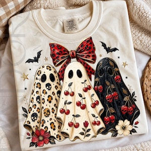 以下が含まれることがあります： クリーム色のTシャツで、3体の幽霊が描かれたグラフィックプリントが施されています。左の幽霊はヒョウ柄にドクロ、中央はサクランボ柄、右は黒色でサクランボとドクロが描かれています。上部には赤いヒョウ柄のリボンがあります。