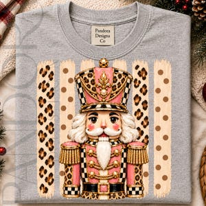 Può includere: Una t-shirt grigio melange con la grafica di uno schiaccianoci nei toni del rosa, nero e oro. Lo schiaccianoci indossa un cappello e dettagli dell'uniforme leopardati, su strisce verticali con stampa leopardata e pois marroni su fondo crema.