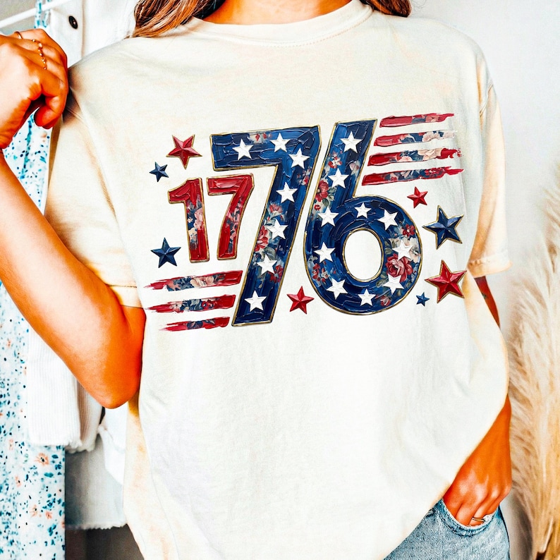 May include: Cream-colored t-shirt featuring the number "76" in blue with white stars and floral accents, and the number "17" in red with floral accents. Red and blue stripes and stars complete the patriotic design.