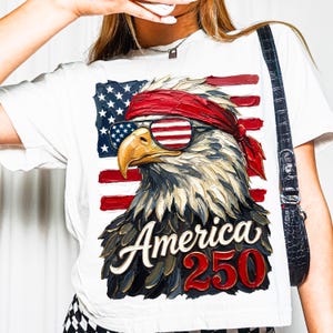 Puede incluir: Camiseta blanca con un dise&ntilde;o patri&oacute;tico de &aacute;guila. El &aacute;guila lleva gafas de sol con el dise&ntilde;o de la bandera estadounidense y una bandana roja. El texto "America 250" est&aacute; en rojo y blanco.
