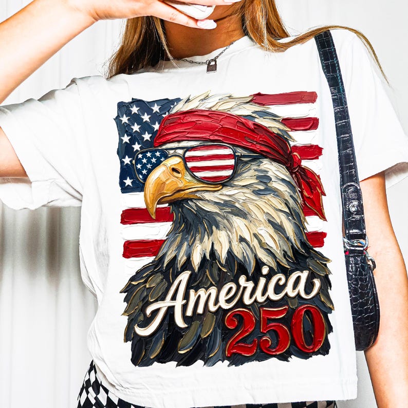 Puede incluir: Camiseta blanca con un dise&ntilde;o patri&oacute;tico de &aacute;guila. El &aacute;guila lleva gafas de sol con el dise&ntilde;o de la bandera estadounidense y una bandana roja. El texto "America 250" est&aacute; en rojo y blanco.