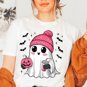 May include: White t-shirt featuring a cartoon ghost wearing a pink beanie and holding a pumpkin and a cup. The ghost has large eyes and a smile. The shirt also has bats and stars printed on it.