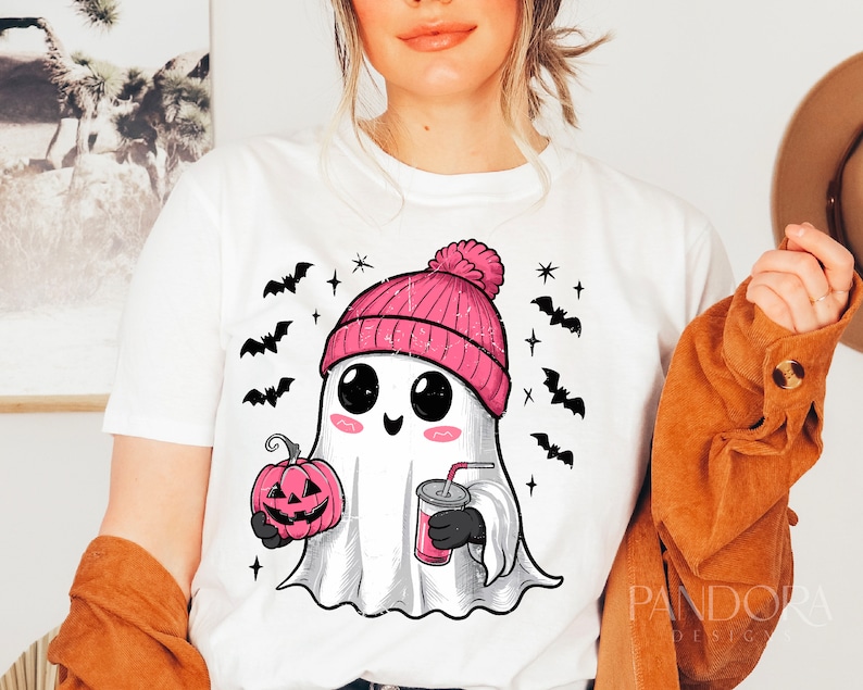 May include: White t-shirt featuring a cartoon ghost wearing a pink beanie and holding a pumpkin and a cup. The ghost has large eyes and a smile. The shirt also has bats and stars printed on it.