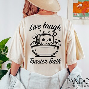 K&ouml;nnte beinhalten: Ein beiges T-Shirt mit einer schwarzen Grafik eines l&auml;chelnden Toasters in einer Badewanne mit Blasen. Der Text "Live laugh Toaster Bath" ist auf das T-Shirt gedruckt.