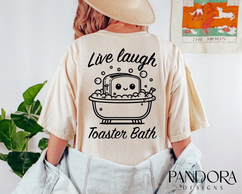 K&ouml;nnte beinhalten: Ein beiges T-Shirt mit einer schwarzen Grafik eines l&auml;chelnden Toasters in einer Badewanne mit Blasen. Der Text "Live laugh Toaster Bath" ist auf das T-Shirt gedruckt.