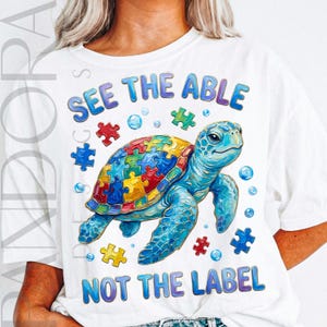 Può includere: T-shirt bianca con un disegno di tartaruga marina blu. Il guscio della tartaruga è un puzzle colorato. La maglietta ha il testo "SEE THE ABLE" e "NOT THE LABEL" in blu e viola.
