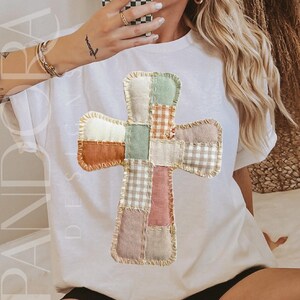 Peut inclure: T-shirt blanc avec un motif de croix en patchwork dans des tons orange, vert et rose. La croix est compos&eacute;e de diff&eacute;rents motifs et textures de tissu. Le t-shirt porte l'inscription "PANDORA DESIGNS".