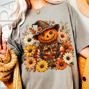 May include: A grey t-shirt featuring a graphic of a scarecrow with a pumpkin head, wearing a hat and a plaid shirt, surrounded by autumn-colored daisies and leaves. The scarecrow has button eyes and a stitched mouth, set against a neutral background.