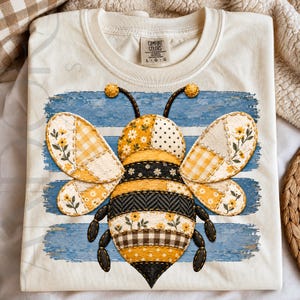 May include: A cream-colored t-shirt featuring a large, central graphic of a bee designed with a patchwork aesthetic. The bee has wings and a body composed of various yellow, white, and black patterns, including florals, gingham, and polka dots.