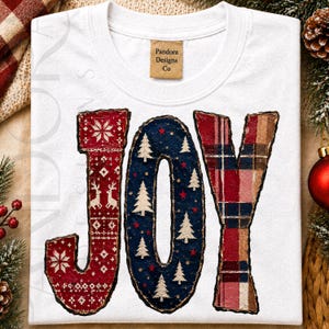 May include: A white short-sleeve t-shirt featuring the word JOY in large letters. The J has a red Nordic pattern with reindeer, the O is dark blue with white pine trees and red stars, and the Y has a red and brown plaid pattern.