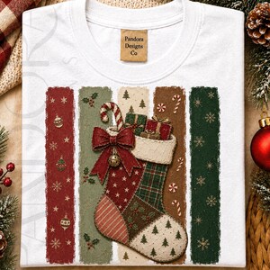 May include: A white crew neck t-shirt featuring a graphic of a Christmas stocking filled with gifts and a candy cane. The stocking is decorated with a red bow and bell, set against five vertical textured stripes in red, green, and beige.