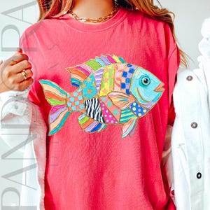 May include: A coral-colored t-shirt featuring a colorful, patchwork-style fish design. The fish has a blue eye and a variety of patterns, including stars, polka dots, and stripes, in shades of blue, green, yellow, and pink. The shirt has a crew neck.