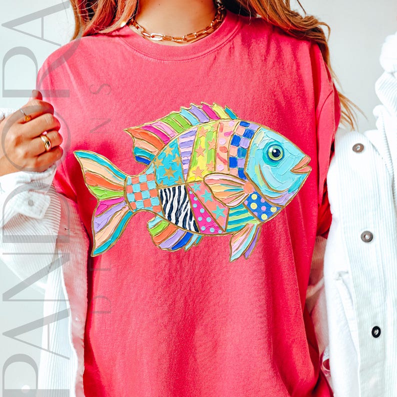 May include: A coral-colored t-shirt featuring a colorful, patchwork-style fish design. The fish has a blue eye and a variety of patterns, including stars, polka dots, and stripes, in shades of blue, green, yellow, and pink. The shirt has a crew neck.