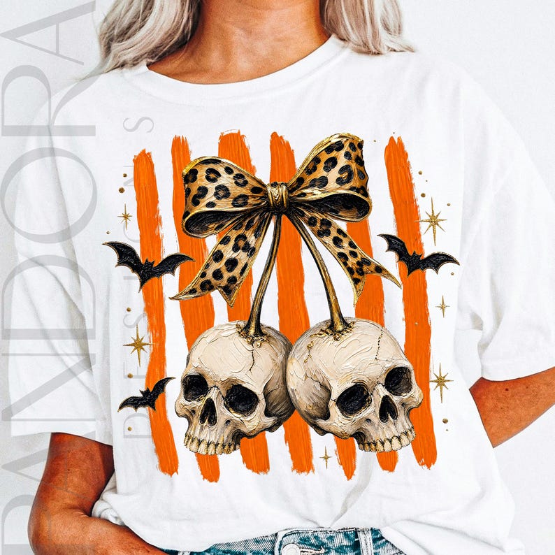 May include: A white t-shirt featuring a graphic of two human skulls hanging from a leopard print bow. The design includes vertical orange brushstroke stripes in the background, small black bats, and gold star accents surrounding the central image.