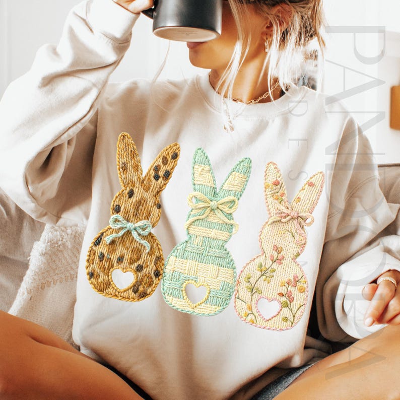 May include: Cream-colored sweatshirt featuring three embroidered bunny designs. The bunnies are in shades of brown, green, and pink, each with a heart-shaped cutout and a bow. The design is perfect for Easter or spring.