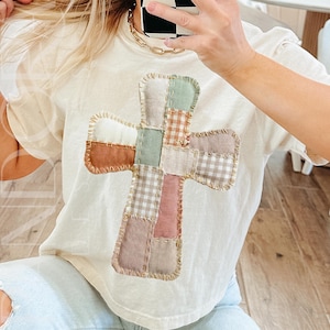 May include: Cream-colored t-shirt featuring a patchwork cross design. The cross is made of various fabric squares in shades of brown, pink, green, and white gingham. The shirt has short sleeves and a relaxed fit, suitable for casual wear.