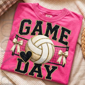 May include: A bright pink t-shirt featuring a graphic print with the text GAME DAY in black block letters. The design includes a white and tan volleyball, a black heart, a black volleyball net, and two cream-colored bows on either side.