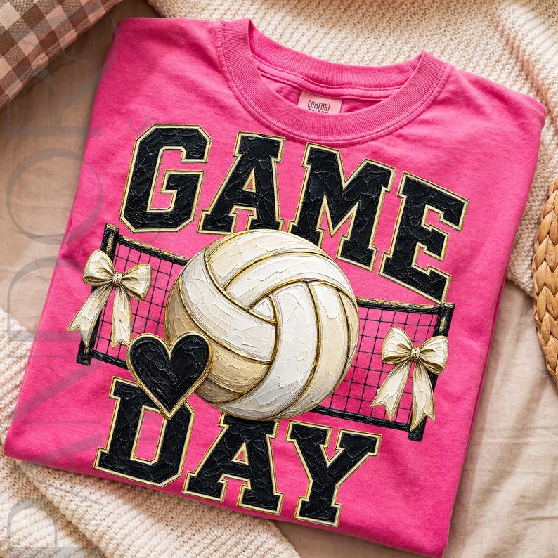 May include: A bright pink t-shirt featuring a graphic print with the text GAME DAY in black block letters. The design includes a white and tan volleyball, a black heart, a black volleyball net, and two cream-colored bows on either side.