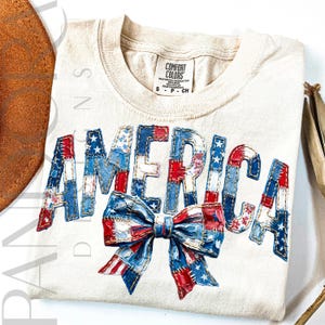 May include: Cream-colored t-shirt with the word "AMERICA" in red, white, and blue patchwork letters. A matching bow is below the word. The shirt is folded and has a tag that says "Comfort Colors".