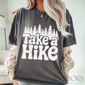 Może przedstawiać: Szary T-shirt z białym nadrukiem, na kt&oacute;rym widnieje napis "Take a Hike" z linią drzew nad tekstem.