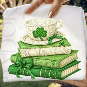 May include: A stack of green books with a white and green knitted teacup and saucer on top. The teacup features a green shamrock. A green bow adorns the books. The image is held by a person.