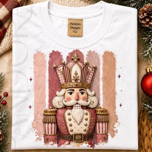 May include: A white t-shirt featuring a graphic of a nutcracker wearing a gold crown and a pink and tan uniform. The design is set against vertical brushstroke stripes in shades of pink and beige. The neck label reads Pandora Designs Co.