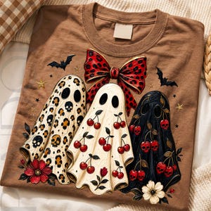 Puede incluir: Una camiseta marr&oacute;n con un gr&aacute;fico de tres fantasmas. El fantasma de la izquierda tiene estampado de leopardo con calaveras, el central tiene cerezas y el de la derecha es negro con cerezas de calavera rojas. Un gran lazo de leopardo rojo corona el dise&ntilde;o.