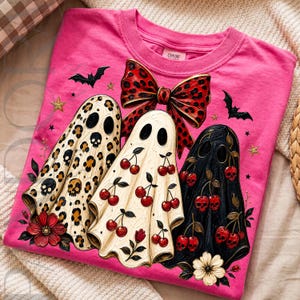 Puede incluir: Una camiseta rosa brillante con un gr&aacute;fico de tres fantasmas. Un fantasma tiene estampado de leopardo con calaveras, otro tiene un patr&oacute;n de cerezas y el tercero es negro con cerezas-calavera. Un gran lazo rojo de leopardo los corona.