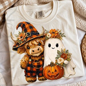 Può includere: T-shirt Comfort Colors color crema con grafica a tema Halloween. La grafica presenta una mucca delle Highlands con cappello da strega e tuta, e un fantasma con una zucca. La mucca e il fantasma sono adornati da elementi floreali.