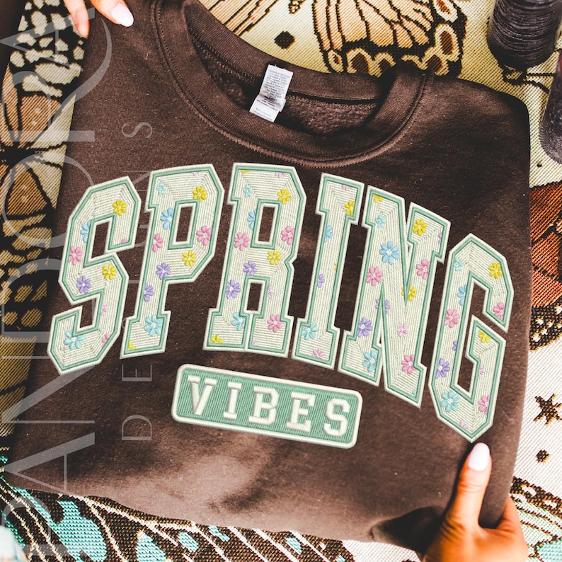 This digital design features the words 'SPRING VIBES' in large, varsity-style block letters. Each letter is filled with small pastel flowers to create a soft, floral pattern. The clean outline gives the lettering a preppy, collegiate look. The design is cheerful, fresh and modern, and is ideal for spring-themed apparel and décor.