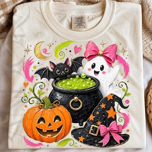 May include: A cream-colored t-shirt featuring a graphic print of a black bat, a white ghost with a pink bow, a black cauldron filled with green liquid, and an orange jack-o'-lantern next to a black witch hat with orange stars and a pink bow.