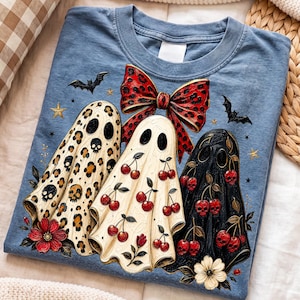 Puede incluir: Una camiseta azul con un gr&aacute;fico de tres fantasmas. El fantasma de la izquierda tiene estampado de leopardo con calaveras, el central tiene un patr&oacute;n de cerezas y el de la derecha es negro con calaveras en forma de cereza. Un lazo rojo arriba.