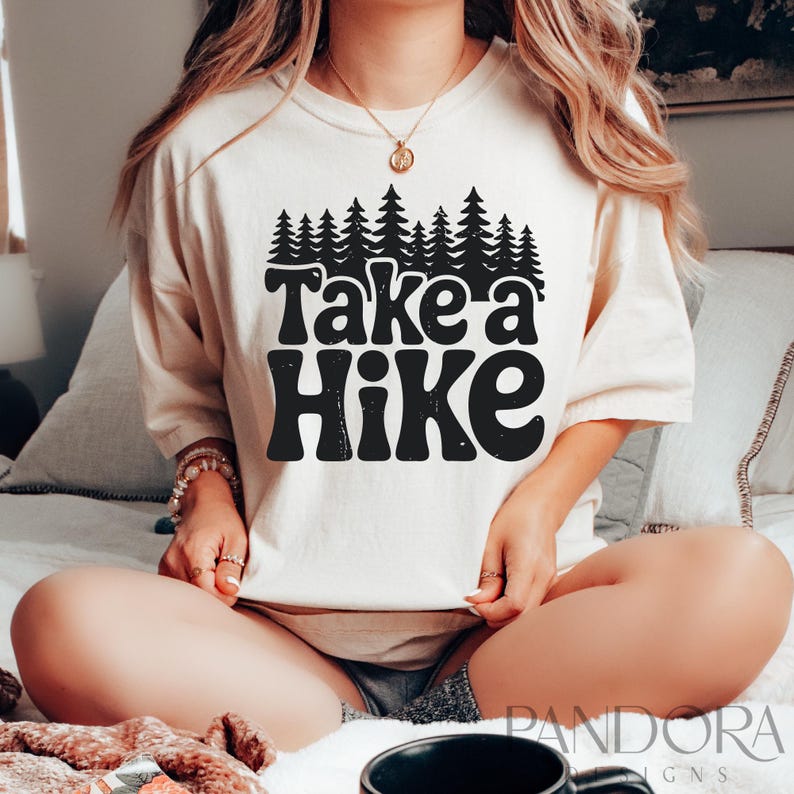 Może przedstawiać: Biały t-shirt z czarnym nadrukiem graficznym, na kt&oacute;rym widnieje napis "Take a Hike" z rzędem drzew nad tekstem. Koszulkę nosi osoba siedząca na białym ł&oacute;żku.
