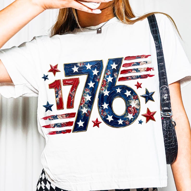 May include: White t-shirt featuring the number "76" in a patriotic design. The "76" is in blue with white stars and floral accents, with red and white stripes. Red and blue stars surround the graphic.