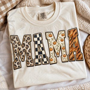 May include: A cream-coloured t-shirt featuring the word MAMA in large block letters. Each letter has a unique pattern: leopard print, black and white checkerboard, a vine print with small orange pumpkins, and a brown and black tiger stripe pattern.