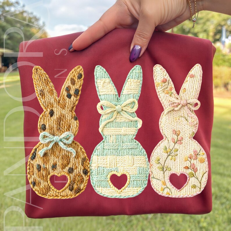 May include: A maroon-colored shirt featuring three embroidered bunny designs. The bunnies have heart-shaped cutouts and are decorated with various patterns, including a speckled design, a striped pattern, and floral embroidery.