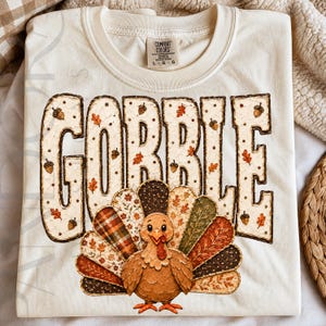 May include: A cream-colored t-shirt featuring the word GOBBLE in large, textured, off-white letters decorated with small brown acorns and orange leaves. Below the text is a cartoon turkey with a fanned tail of patterned, multi-colored feathers.