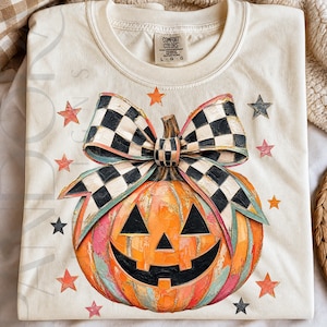 May include: A cream-colored t-shirt featuring a large, textured orange jack-o'-lantern with a black and white checkered bow on top. The design is surrounded by small orange and black stars. The shirt has a Comfort Colors brand label at the neck.
