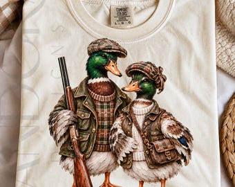 May include: A cream-colored t-shirt featuring a graphic of two mallard ducks dressed in outdoor clothing. The left duck wears a plaid sweater, green jacket, and flat cap while holding a shotgun. The right duck wears a sweater, vest, and cap with a bag.